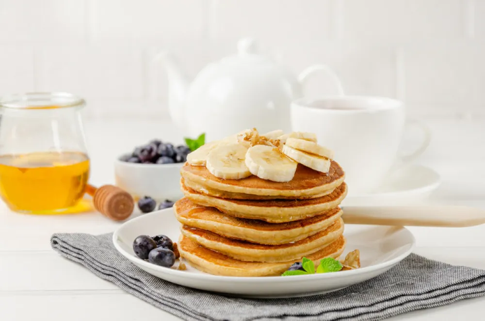A stack of oatmeal banana pancakes with slices of fresh bananas, walnuts and honey on top with cup of tea on a white wooden background. A healthy breakfast. Copy space