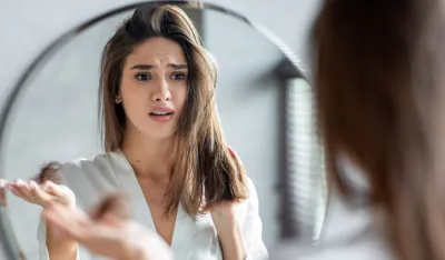 Portrait of stressed young woman with bunch of fallen hair in hand looking at mirror in bathroom, scared upset millennial lady in white silk robe suffering hairloss problem, selective focus