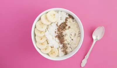 Milk oatmeal with flax seeds, banana and coconut chips in the white bowl in the center of the pink background. Copy space. Top view.