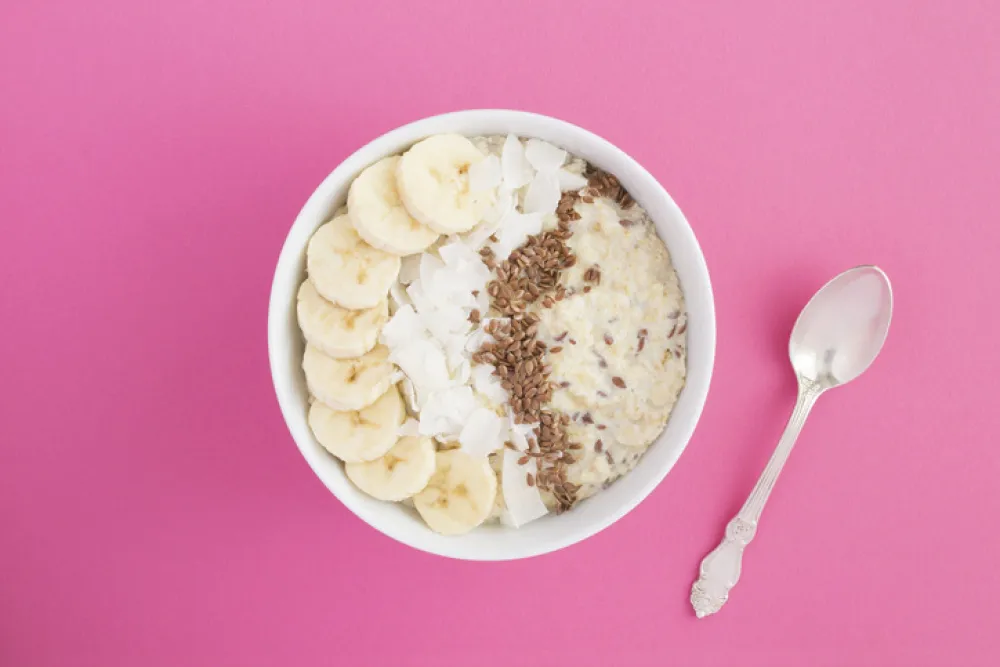 Milk oatmeal with flax seeds, banana and coconut chips in the white bowl in the center of the pink background. Copy space. Top view.