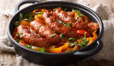 Grilled homemade sausages with colored peppers, onions, herbs and tomatoes closeup in a frying pan on the table. horizontal<br>