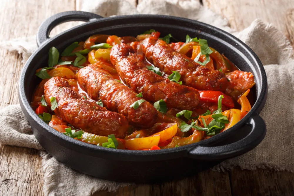 Grilled homemade sausages with colored peppers, onions, herbs and tomatoes closeup in a frying pan on the table. horizontal<br>