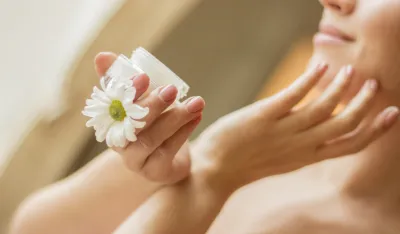 Spring portrait of gentle pretty girl looking away and smiling while applying moisturizing lotion on face. Beautiful young woman holding jar of cream in hand. Gentle girl, skin or body care, treatment