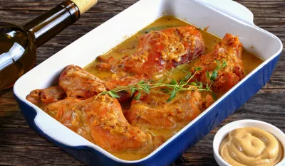 appetizing Rabbit Cooked in mustard sauce or lapin a la moutarde in gratin dish on wooden table with bottle of wine and garlic on background, authentic french recipe, view from above, close-up