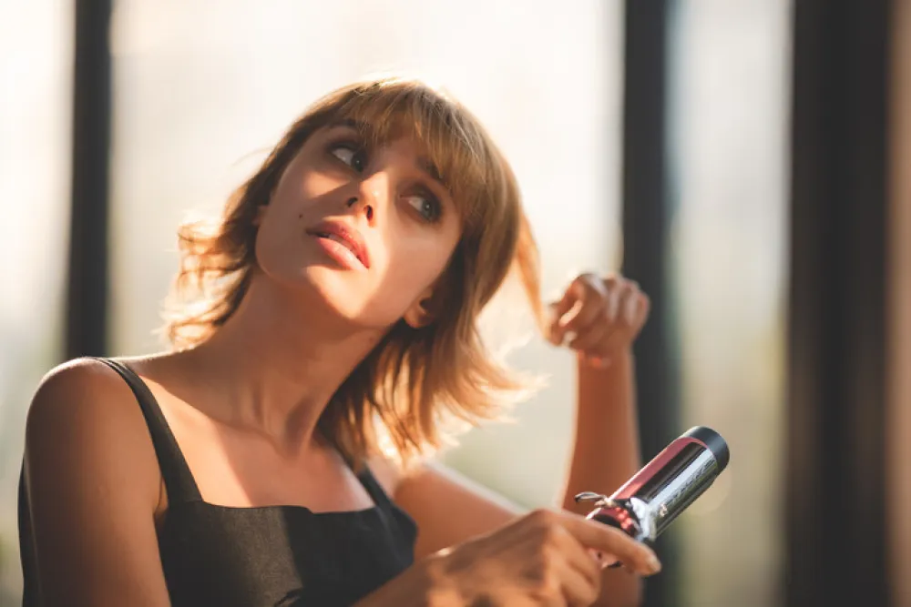 Young beautiful blonde woman with short hair using electronic curling iron styling her hair for fashion and trying new lifestyle at home by looking at self in mirror