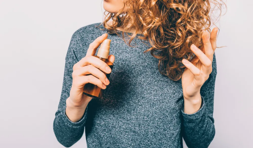 Young woman holding bottle with nutritional oil applying on her curly brown hair. Female's hands using cosmetic serum to prevent split ends.
