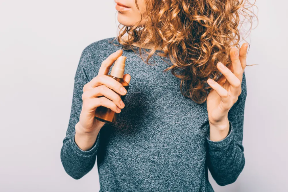 Young woman holding bottle with nutritional oil applying on her curly brown hair. Female's hands using cosmetic serum to prevent split ends.