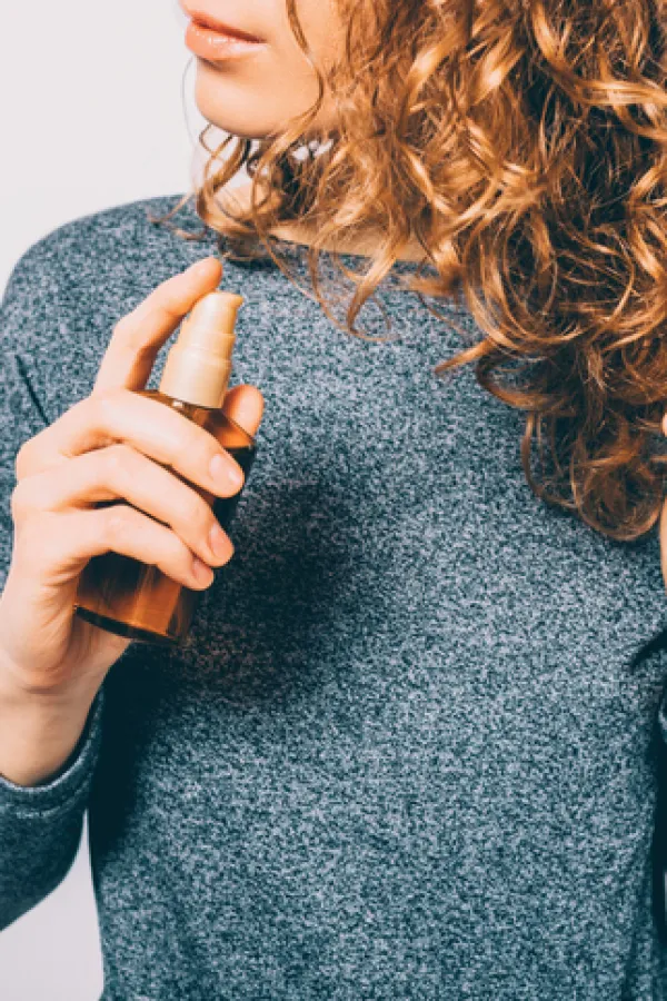 Young woman holding bottle with nutritional oil applying on her curly brown hair. Female's hands using cosmetic serum to prevent split ends.