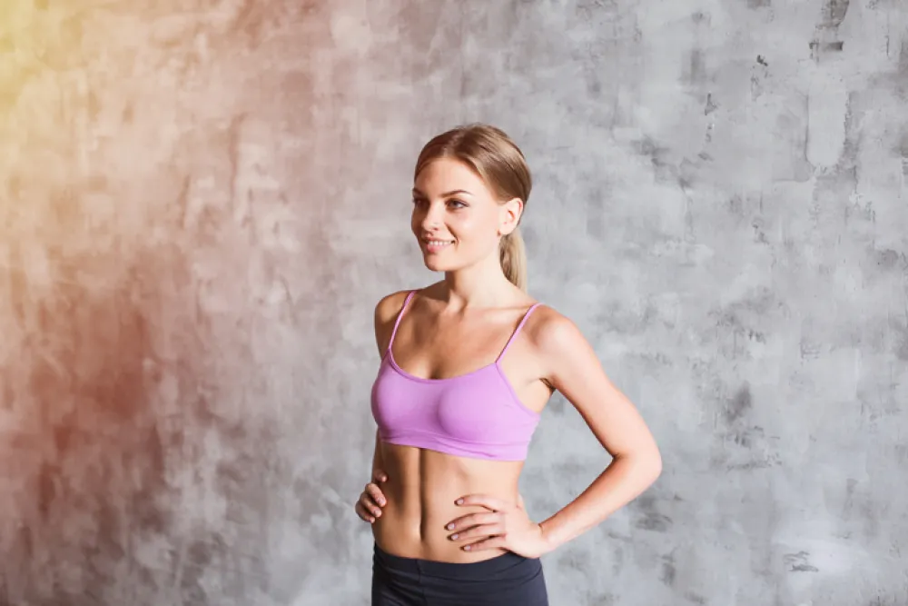 Young pretty woman dressed in a pink top with her hands on hips in the grey background sunlight effect