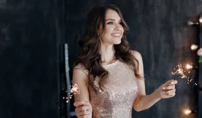Girl with a sparkler near the Christmas tree. A cheerful young woman with a cute smile in a beige dress is standing and holding a sparkling sparkler in the hands against the background of the Christmas tree. Beautiful girl celebrates Christmas.