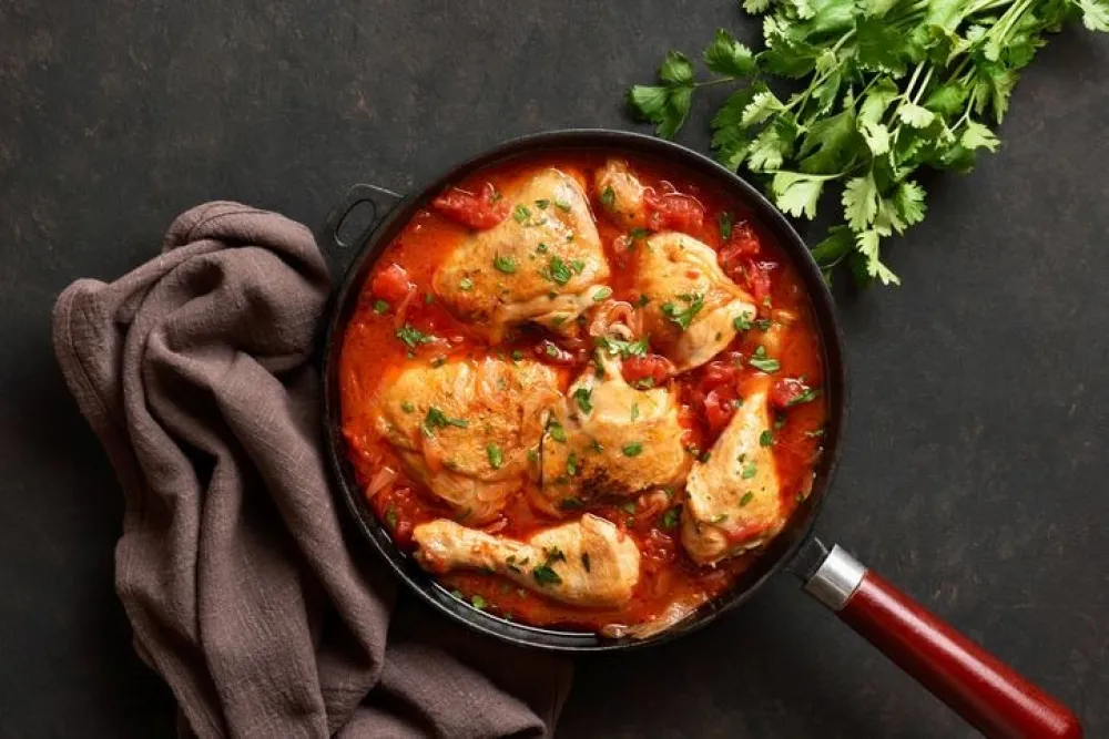 Georgian chicken stew with tomatoes and herbs (chakhokhbili) in cast iron skillet over dark stone background. Baked chicken with tomato vegetable gravy. Top view, flat lay
