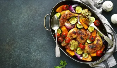 Chicken legs roasted with vegetables in a skillet pan over dark slate,stone or concrete background.Top view.