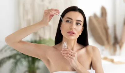 Modern beauty products at home, cosmetology, anti-aging cosmetics and advice from blogger and specialist. Young brunette lady in towel applies oil to skin of face, on bedroom interior background